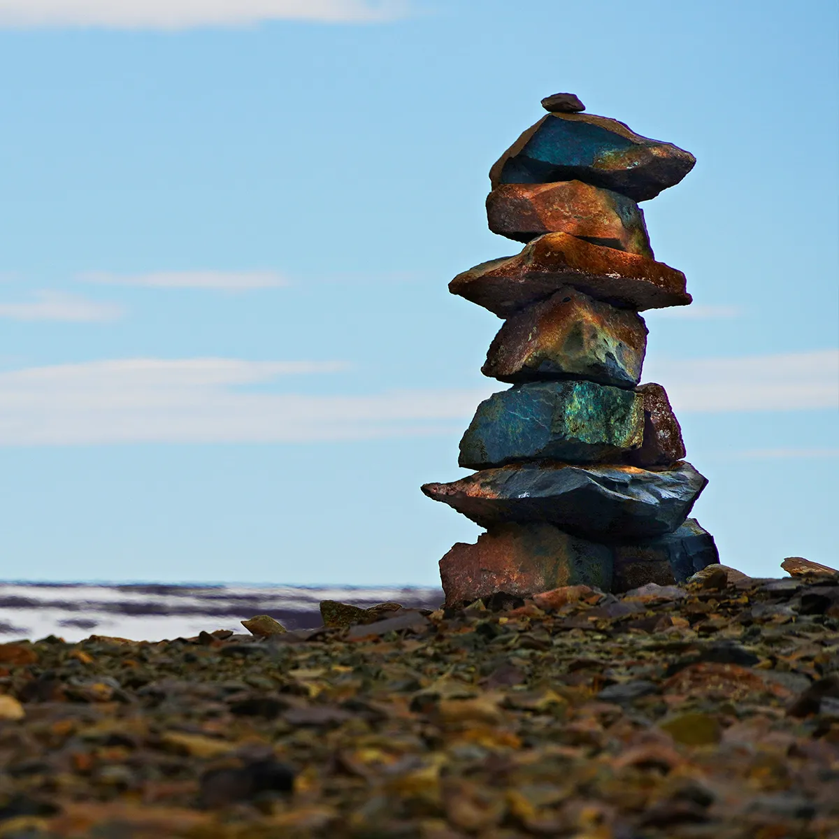 Stack of flat stones balanced on rocky ground with a blue sky and distant water in the background.