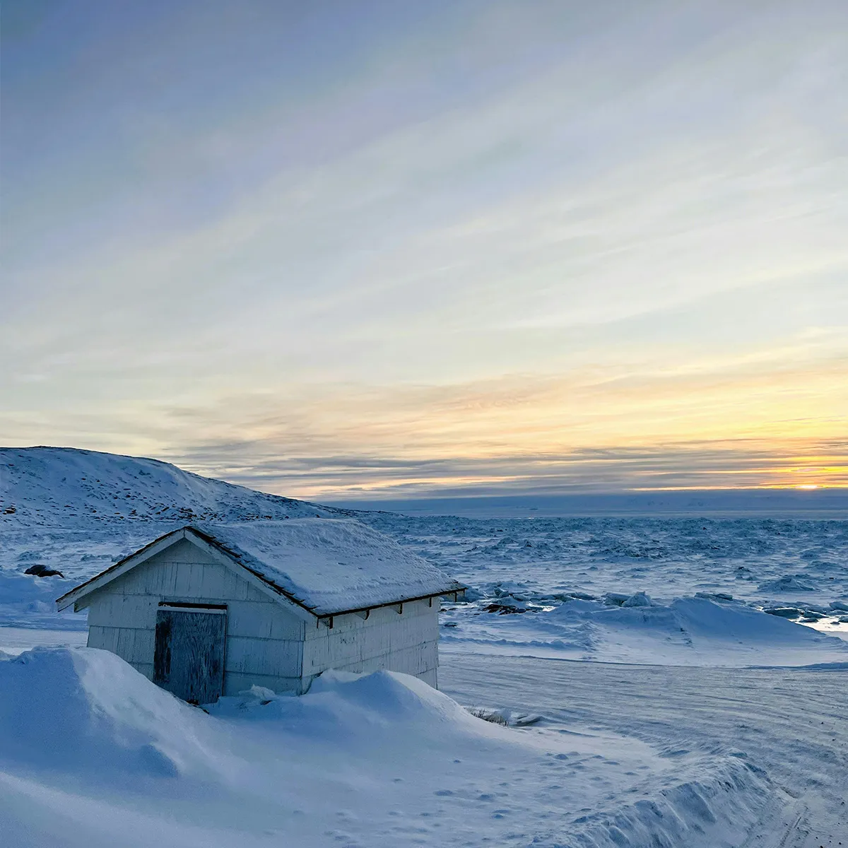 A small white shed sits in a snowy, icy landscape near a frozen sea under a pale sunset sky.