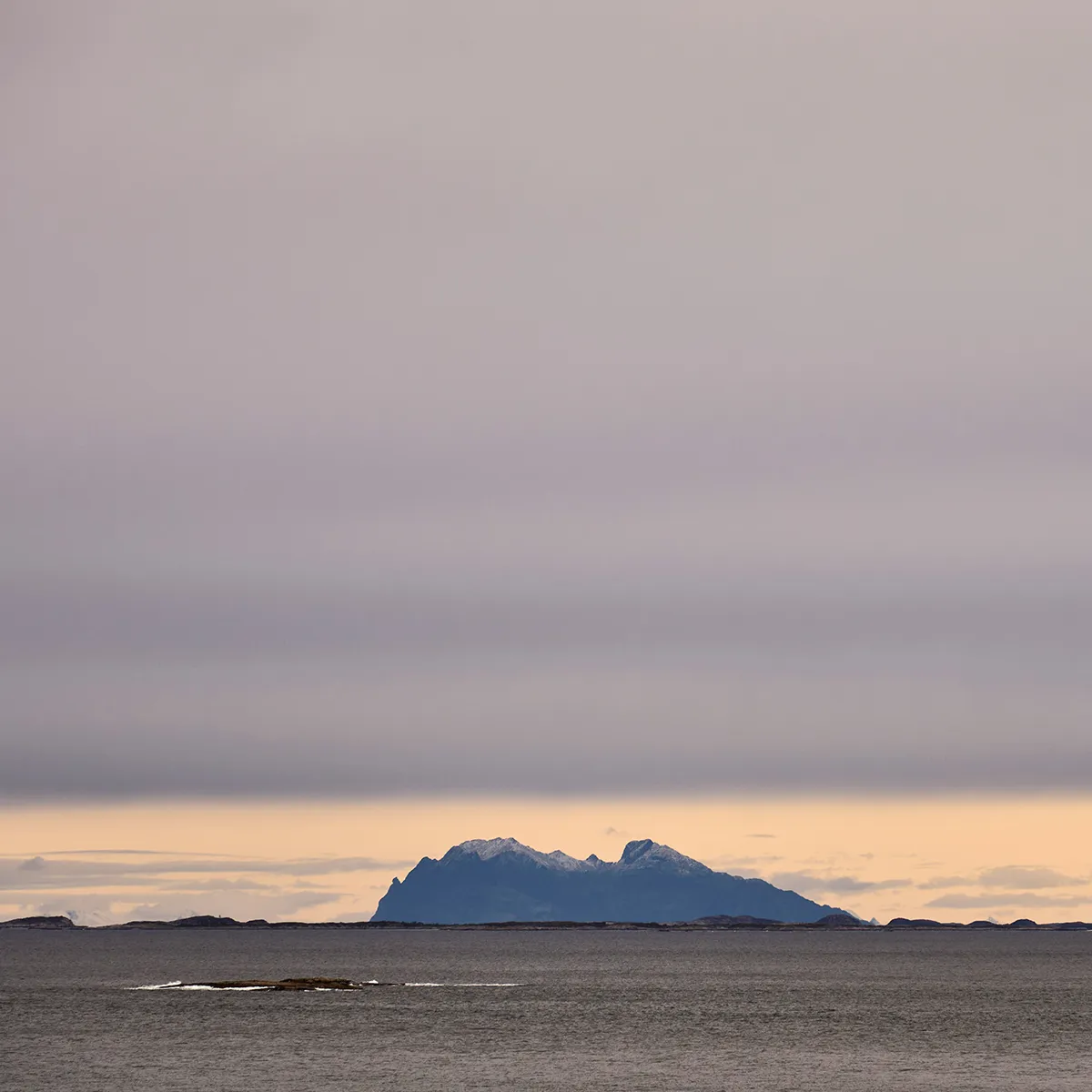 Distant snowy mountains under a cloudy sky, seen across calm water at sunset.