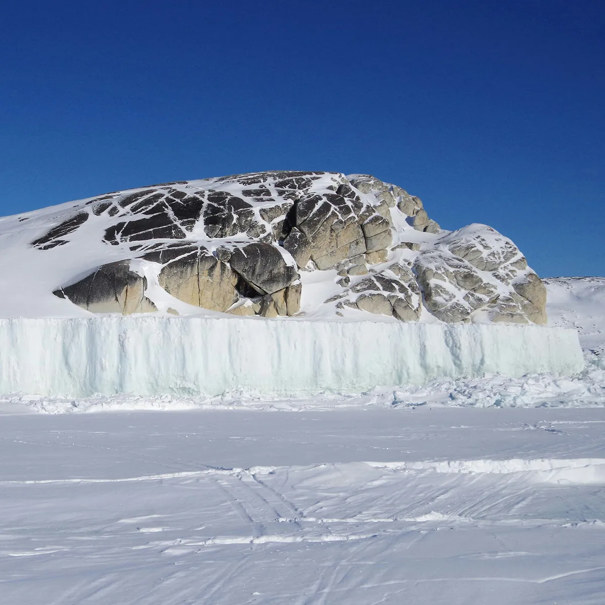 Snow-covered rocky hill with a wall of ice below, under a clear blue sky in a frozen landscape.
