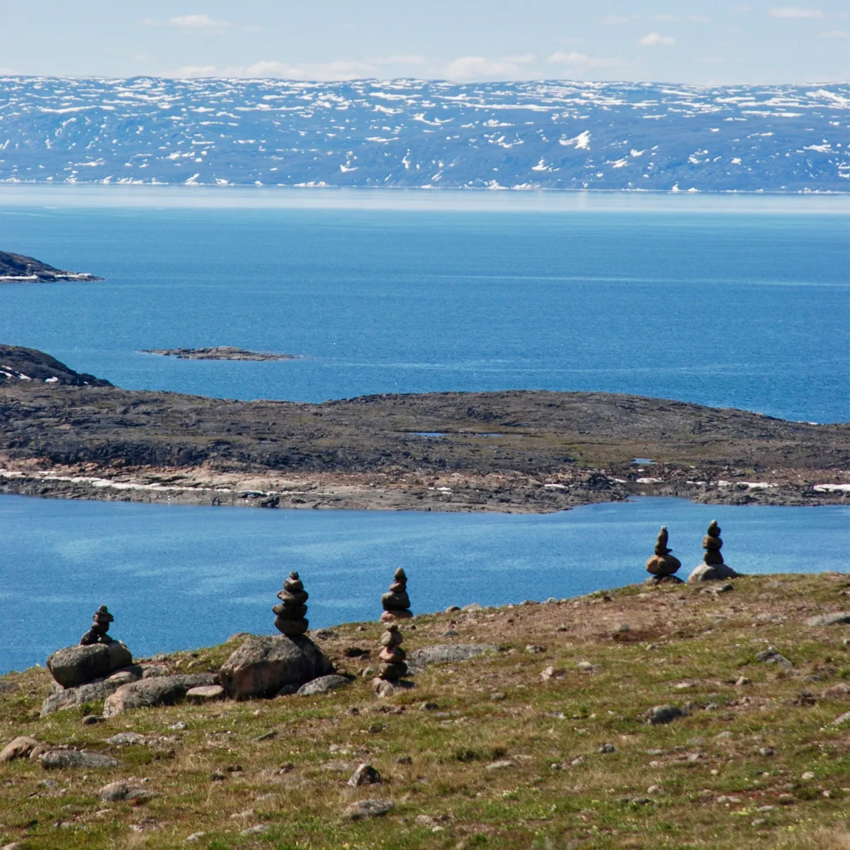 Stone cairns on grassy hill overlook blue water and snow-dotted mountains under a partly cloudy sky.
