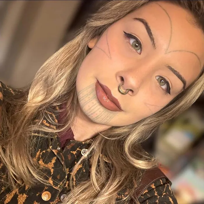 A young woman with face markings, nose ring, and long blonde hair, wearing a patterned jacket, smiles softly.