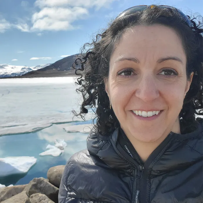 Smiling woman in a puffy jacket stands by a snowy, frozen lake with mountains in the background.