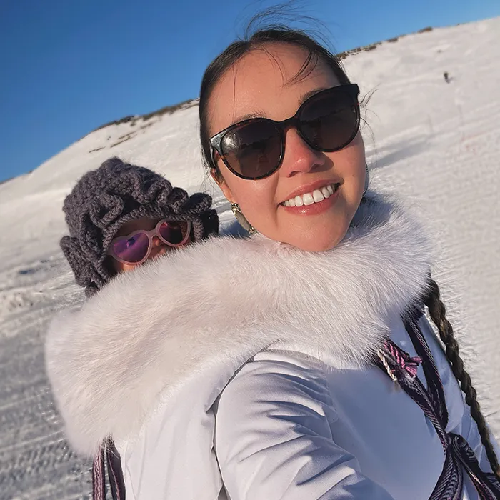 A mother carrying a baby in winter clothes smiling for a selfie on a sunny snowy landscape.