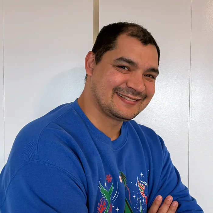 Man smiling in a blue sweater with arms crossed, standing in front of a white background.