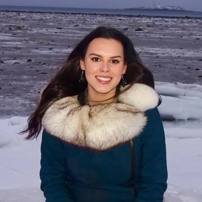 Woman in a blue coat with a fur collar smiling, sitting outdoors in a snowy, icy landscape.