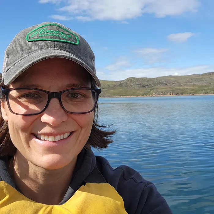 Woman wearing glasses and a cap smiling by a calm lake with hills and blue sky in the background.