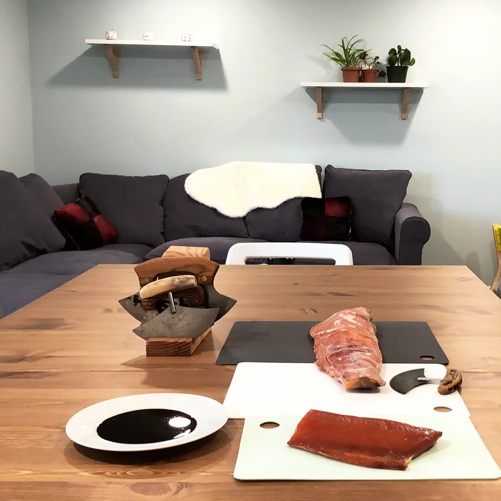 A wooden table with fish fillets, a cutting board, knife, and a plate of dark sauce in a cozy living room.